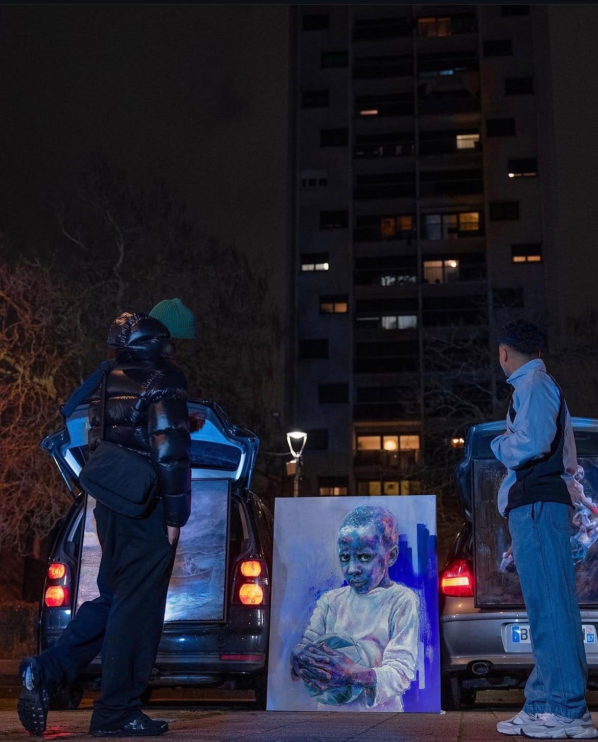La nuit, au pied d'une tour d'habitation éclairée, deux voitures au coffre ouvert, des tableaux posés dans les coffres ; entre elles, un grand portrait d'enfant peint aux tons bleus est posé au sol, et deux personnes se tiennent face aux coffres.